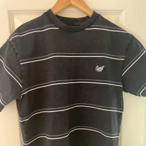 Svart randig t-shirt från Sweet Skies - Svart t-shirt med tunna vita och blå horisontella ränder från Sweet Skies. Klassisk passform med rund halsringning och broderad logga på bröstet. Tillverkad i 100% bomull för en mjuk och skön känsla. Storkel S men passar som M