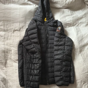 Svart pufferjacka från Parajumpers - Säljer en svart pufferjacka från Parajumpers med huva och dragkedja. Jackan är lättviktig och har ett broderat märke på ärmen. Perfekt för kyliga dagar och enkel att bära med sig. Jackan har 1 hål men är lagat så de syns inte så mycket men de ser ut som på bilden annars inga andra skador