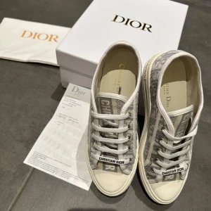Dior skor - Säljer ett par gråa Dior sneakers med tydligt Dior-mönster och vit sula. Skorna har rund tå och platt sula, samt är tillverkade i textil med detaljer i syntet. Klassisk och stilren design med Dior-logga på hälen. Helt nya bara testade! De är i storlek 40 hör av er vid frågor!