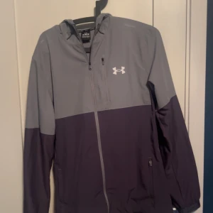 Grå och svart vindjacka från Under Armour - Snygg tvåfärgad vindjacka från Under Armour i grått och svart. Jackan har huva, dragkedja framtill och sidofickor med dragkedja. Perfekt för träning eller när du vill ha en sportig look.