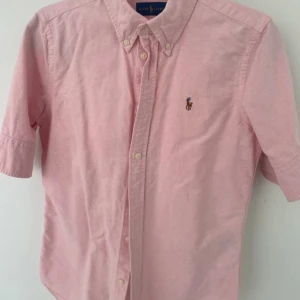 Rosa linne piké från Ralph Lauren - Säljer en klassisk rosa lille piké från Ralph Lauren med korta ärmar och button-down krage. Skjortan har broderad logga på bröstet och stängs med knappar framtill. Perfekt för en stilren och avslappnad look.