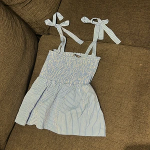 Blåvit randig kortklänning med rosettband - Supersöt kortklänning i blåvitt randigt med smockad överdel och knytbara axelband i rosett. Perfekt för varma dagar och ger en fräsch sommarkänsla. Lätt och luftig modell som passar till många tillfällen.