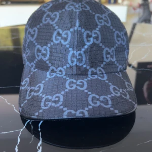 Svart keps från Gucci med GG-mönster - Snygg svart keps från Gucci med det klassiska GG-mönstret i grått över hela kepsen. Justerbart spänne baktill i läder och tyg i polyester. Perfekt accessoar för att lyfta din stil.