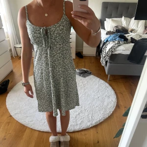 Grön blommig klänning med knyt framtill - Söt grön klänning med vita blommor och smala axelband. Klänningen har knyt framtill vid bysten och en lätt utställd passform som slutar ovanför knäna. Endast använd ett fåtal gånger. Klänning är från lager 157 och är i storlek S