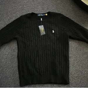 Svart kabelstickad tröja från Polo Ralph Lauren - Kabelstickad svart tröja från Polo Ralph Lauren med rund halsringning och broderad logga på bröstet. Klassisk och stilren modell med långa ärmar och ribbade muddar. Perfekt för lager-på-lager eller som den är pris kan diskuteras vid snabb affär 