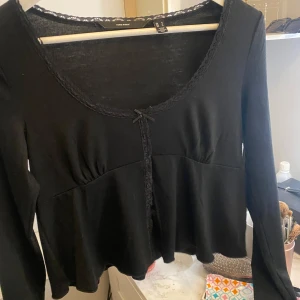 Svart långärmad topp med spets från Vero Moda - Säljer en svart långärmad topp från Vero Moda med spetsdetaljer längs urringningen och en liten rosett framtill. Modellen är lätt utsvängd nertill. Aldrig använd