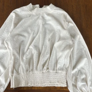 Sommrig beige blus från Grunt - Fin sommrig blus från Grunt med fina blomdetaljer och resår längst ner för att ge volym. Blusen stängs med knappar i nacken och passar perfekt för en fin sommarkväll.