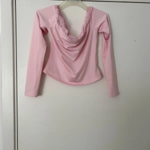 Ljusrosa långärmad topp med spets - Säljer en ljusrosa långärmad topp med vacker spetsdetalj runt den djupa ringningen. Toppen har en mjuk, draperad framsida och är tillverkad i ett stretchigt material som sitter snyggt på kroppen.