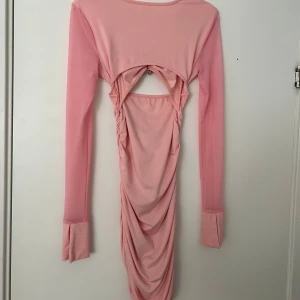 Rosa långärmad klänning med hjärtformad detalj - Trendig rosa klänning med långa ärmar och draperad kjol. Framtill finns en hjärtformad detalj med pärlor och en liten cut-out under bysten. Klänningen har en tight passform och är tillverkad i ett stretchigt material.