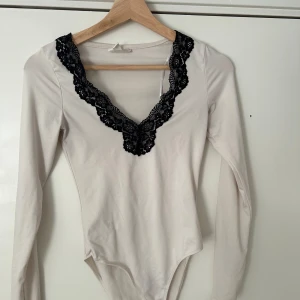 Vit långärmad body med svart spets - Säljer en vit långärmad body med djup v-ringning dekorerad med svart spets. Perfekt att styla till både jeans och kjol för en trendig look. Mjuk och stretchig i materialet.