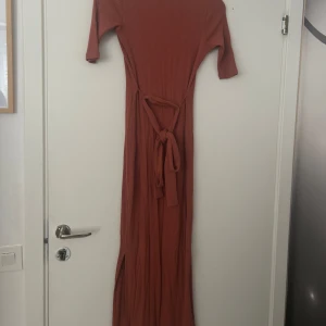 Rostbrun ribbad långklänning med knappar - Säljer en långklänning i rostbrun färg med ribbad struktur. Klänningen har v-ringning, knappar hela vägen framtill, trekvartsärm och ett matchande bälte i midjan. Perfekt för dig som gillar en stilren och bekväm look har även en två slits.