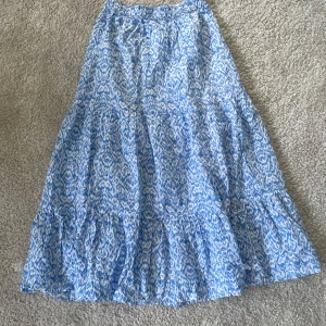 Blåvit mönstrad långkjol från Lindex - Säljer en luftig blåvit långkjol från Lindex med elastisk midja och ett snyggt abstrakt mönster. Kjolen har volangdetaljer och är perfekt för varma dagar. Superfin att styla med en enkel topp eller linne.