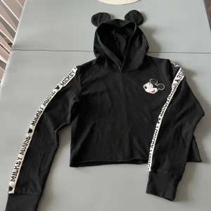 Svart croppad hoodie med Musse Pigg från H&M x Disney - Svart croppad hoodie med öron på huvan och Musse Pigg-tryck på bröstet. Band med texten 'Mickey Mouse' längs ärmarna. Mjuk insida och långa ärmar med muddar. Perfekt för dig som gillar Disney och streetstyle. Liten fläck på ena tröjärmen- se bild