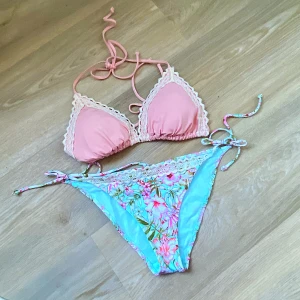 Rosa och blommig bikini från H&M - Superfin bikini från H&M med rosa trekantsöverdel och blommigt mönstrade trosor i blått och rosa. Båda delarna har vita spetsdetaljer och knytband. Perfekt för sommaren och strandhäng! Trosan är ny med lappar 