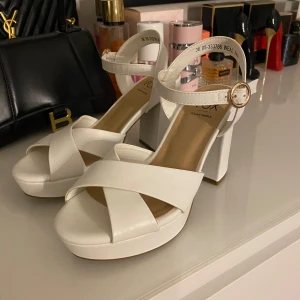 Vita platåsandaler med klack från VOX - Säljer ett par vita sandaler från VOX med bred klack och platåsula. Skorna har öppen tå, korsade remmar framtill och justerbart spänne med gulddetalj vid ankeln. Använda 1 gång, Ny pris 449🌸