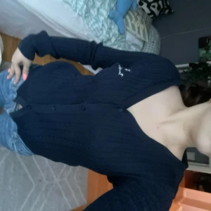 Mörkblå kabelstickad kofta från Bondelid  - Säljer en mörkblå kabelstickad kofta från Boomerang med broderad logga på bröstet. Koftan har v-ringning, knappar framtill och långa ärmar. Perfekt att slänga över en t-shirt eller linne.