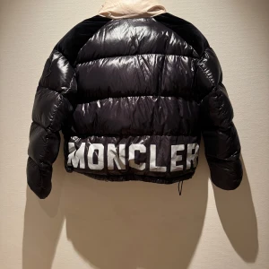 Svart dunjacka från Moncler - Säljer en svart, glansig dunjacka från Moncler med stor vit logga på ryggen och kontrasterande beige krage. Jackan har lång ärm, dragkedja och en klassisk pufferstil. Perfekt för kyliga dagar när du vill ha både värme och stil.