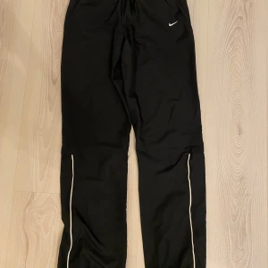 Svarta träningsbyxor från Nike med vita detaljer - Svarta träningsbyxor från Nike med vita ränder längs benen och liten vit logga framtill. Byxorna har dragkedjor vid bensluten och resår i midjan. Tillverkade i 100% polyester, perfekta för sport och aktivitet.