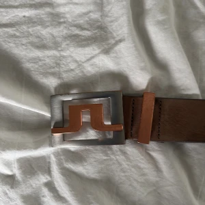 Brunt bälte med unik spänne - Säljer ett brunt bälte i skinn med ett stilrent och unikt spänne i silver och kopparfärg. Bältet har en bredare design och flera hål för justering. Perfekt accessoar för att lyfta din outfit.