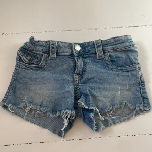 Blå jeansshorts med fransar - Säljer ett par blå jeansshorts med fransig kant och klassiska fickor både fram och bak. Shortsen har bälteshällor, knappstängning och en cool, sliten look. Perfekta för varma dagar och en avslappnad stil.