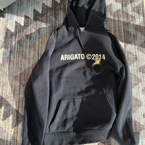 Marinblå hoodie från Axel Arigato - Säljer en marinblå hoodie från Axel Arigato med tryck 'ARIGATO ©2014' och en broderad fågel på bröstet. Klassisk känguruficka framtill och justerbar huva. Perfekt för en avslappnad stil.