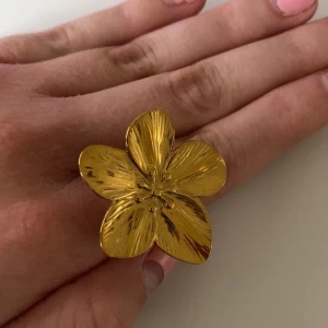 Guldfärgad blomring - Säljer en stor och iögonfallande guldfärgad ring formad som en blomma med fem blad. Ringen har en blank och lätt texturerad yta som ger ett unikt uttryck. Perfekt för dig som vill sticka ut med dina accessoarer!
