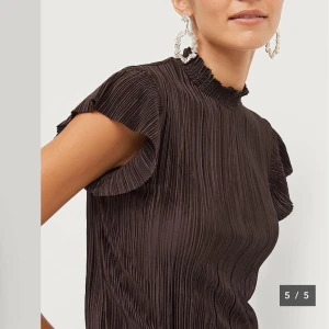 Brun blus från Ellos - Säljer en brun plisserad blus från Ellos med korta volangärmar. Blusen har en elegant struktur och är tillverkad i ett lätt, syntetiskt material. Perfekt för dig som gillar stilrena och trendiga toppar. Helt oanvänd med prislapp kvar. Köpt för 249kr.