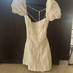 Vit kortklänning med puffärmar och snörning - Superfin vit kortklänning med puffiga ärmar och snygg snörning i ryggen. Klänningen har en feminin passform och är perfekt för sommaren och studenten. Materialet känns lätt och luftigt, och de dekorativa banden ger en extra detalj.