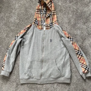 Grå zip-hoodie från Burberry med rutiga detaljer - Grå hoodie med dragkedja från Burberry. Klassiska Burberry-rutiga detaljer på huva och ärmar. Fickor framtill och broderad logga på bröstet. 