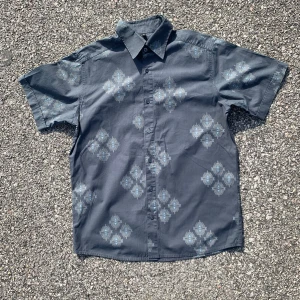 Mörkblå kortärmad skjorta med mönster - Säljer en mörkblå kortärmad skjorta med ljusblå geometriskt mönster. Skjortan har klassisk krage och knäppning framtill. Perfekt för dig som vill ha en stilren men ändå unik look.