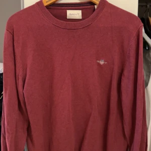 Vinröd tröja från GANT - Vinröd långärmad tröja från GANT med rund halsringning och diskret broderad logga på bröstet. Klassisk och stilren modell som passar perfekt till många olika outfits. 