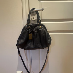 Svart handväska från Marc Jacobs - Säljer en svart handväska i skinn från Jacobs. Väskan har både handtag och avtagbar axelrem i svart, guldfärgade metalldetaljer och dragkedja. Den har några få defekter som visas på bild 5.