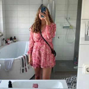 Rosa blommig klänning - Söt kortklänning i rosa med blommigt mönster och volangdetaljer. Klänningen har v-ringning och långa ärmar i ett lätt, luftigt material. Perfekt för varma dagar eller sommarens tillfällen💓Använd 2 gånger förra sommaren men säljer nu pga den är för liten. 