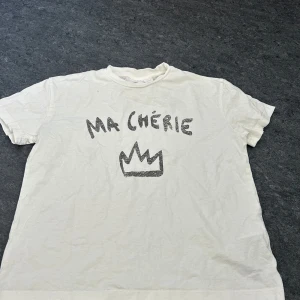 Vit t-shirt från Gina Tricot - Säljer en vit t-shirt från Gina Tricot med texten 'MA CHÉRIE' och en krona tryckt på framsidan. T-shirten har en rund hals och korta ärmar. Perfekt för en avslappnad stil.