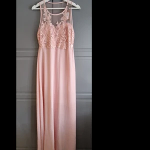 Ljusrosa balklänning med spetsdetaljer - Ljusrosa balklänning/långklänning med spetsdetaljer och genomskinliga paljetter (i spetsmönstret, ger ett skimmer) på överdelen. Diskret dragkedja i sidan. Stl 40/M. Längd 162 cm. Midjebredd på plan yta 38 cm. 