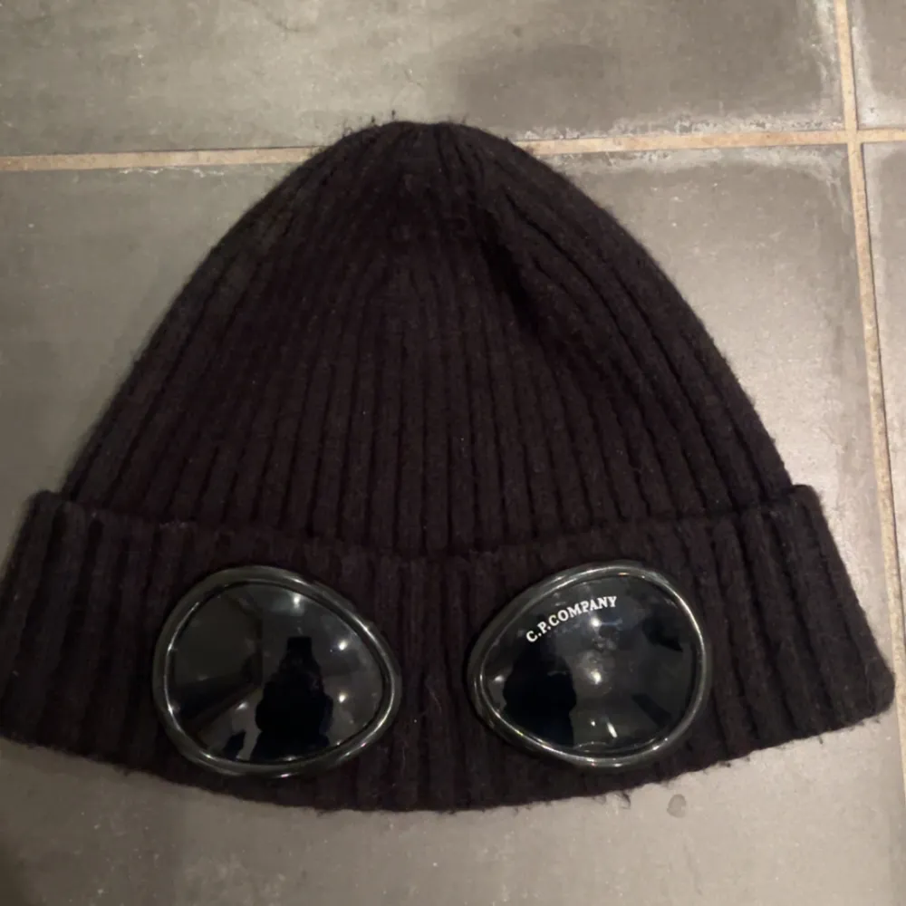 Snygg svart ribbad mössa från C.P. Company med unika glasögondetaljer framtill. Perfekt för att hålla stilen och värmen under kyliga dagar.För fler frågor kom privat.. Asusteet.