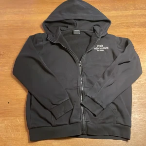 Svart hoodie från Peak Performance - Säljer en svart hoodie från Peak Performance med dragkedja och huva. Den har en broderad logotyp på bröstet och är perfekt för en avslappnad stil. Passar bra till både vardag och träning. Storlek 170 passar som storlek xs och s.