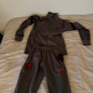 Grå hoodie och byxor från nike/ psg - Säljer en grå hoodie och matchande byxor från Nike. Hoodien har en röd Nike-logga och ett emblem på bröstet. Byxorna har också en röd Nike-logga. Perfekt för träning eller en avslappnad dag.