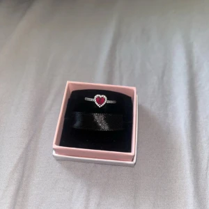 Ring med hjärta från Pandora - Säljer en elegant ring från Pandora med ett rött hjärta i mitten, omgiven av små glittrande stenar. Ringen har en tunn silverfärgad banddesign som ger en stilren look. Perfekt för att ge en touch av romantik till din outfit. Köpt för 899kr