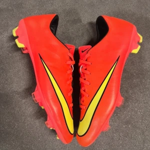 Röda fotbollsskor från Nike - Snygga röda fotbollsskor från Nike med gula och svarta detaljer. Skorna har snörning och är designade för optimal prestanda på planen. Perfekta för dig som vill sticka ut med stil och funktion.