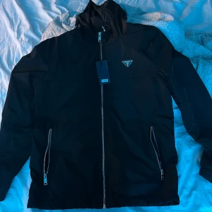 Svart jacka från Prada - Snygg svart jacka från Prada med dragkedja och huva. Jackan har en stilren design med två fickor framtill och en diskret logotyp på bröstet. Perfekt för en modern look. Jackan är aldrig använd då den var för kort i armarna.      OBS!!!!!priset där uppe stämmer inte 