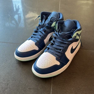 Nike Air Jordans 1 Mint Foam - Snygga Nike Air Jordans i en fräsch kombination av blått och vitt med mintgröna detaljer. Skorna har en klassisk hög design med snörning och den ikoniska Nike-loggan på sidan. Perfekta för dig som vill ha en stilren look med en touch av färg.