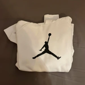 Vit hoodie från Jordan - Säljer en stilren vit hoodie från Jordan med det ikoniska Jumpman-logotypen i svart på framsidan. Tröjan har en klassisk känguruficka och en bekväm huva. Perfekt för en sportig look eller avslappnade dagar.