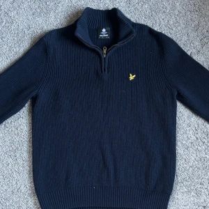 Mörkblå stickad tröja från Lyle & Scott - Säljer en stilren mörkblå stickad tröja från Lyle & Scott med dragkedja vid halsen och ett broderat gult emblem på bröstet. Perfekt för kyligare dagar. Tröjan har långa ärmar och ribbade kanter för en bekväm passform.