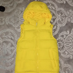 Gul dunväst från McKinley - Säljer en ljusgul dunväst från McKinley med huva och dragkedja. Luvan är tillräcklig tjock för att hålla värmen utan att ha mössa. Perfekt för kyliga dagar när du vill hålla värmen utan att bära en jacka. Västen har en sportig look och är lätt att matcha med andra kläder. Västen passar de som är ca 158-160 cm/13-14 år.
