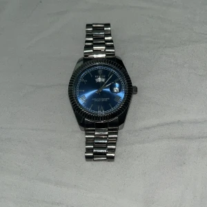 Silverfärgad klocka med blå urtavla - Snygg klocka med en stilren blå urtavla och datumvisning. Armbandet är i silverfärgad metall och justerbart med extra länkar. Perfekt för den som gillar en klassisk look med en modern touch. PRIS KAN DISKUTERAS 