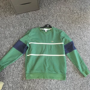 Grön tröja från Lacoste - Säljer en grön tröja från Lacoste med vita ränder och mörkblå detaljer på ärmarna. Bara att fråga om ni har fler frågor👍