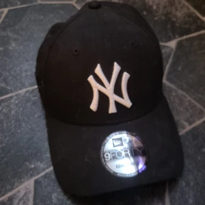 Svart keps med vit logga - Snygg svart keps från New Era med vit broderad logga framtill. Justerbar passform med klassisk design. Perfekt för en casual look. Är använd ungefär 3 gånger men har dock stått ett tag så skicket ser värre ut men egentligen är den nästan ny