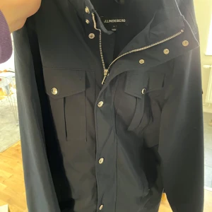 Mörkblå jacka från J.Lindeberg - Snygg blå jacka från J.Lindeberg med dragkedja och knappar framtill. Jackan har två bröstfickor med knappstängning och en stilren design. Perfekt för en modern look.