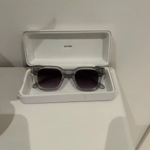 Chimi 04 - Snygga grå solglasögon från Chimi med transparent båge och mörka linser. Perfekta för att ge en cool touch till din stil. Kommer i ett stilrent vitt fodral. Använda ett fåtal gånger